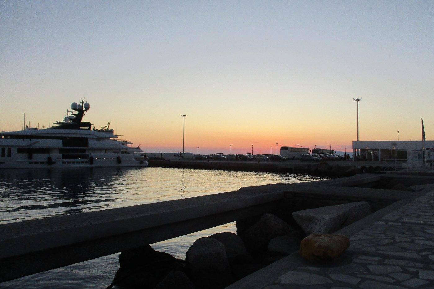 A large yacht moored in the harbour of Mykonos at sunset, with the sky glowing orange over the sea