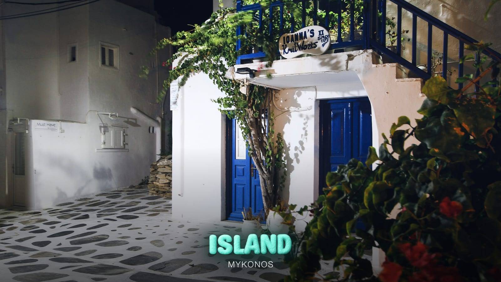 A whitewashed lane of Mykonos Town (Chora) at night, with blue doors, a wooden staircase and bougainvillea, with the Party Island Mykonos logo