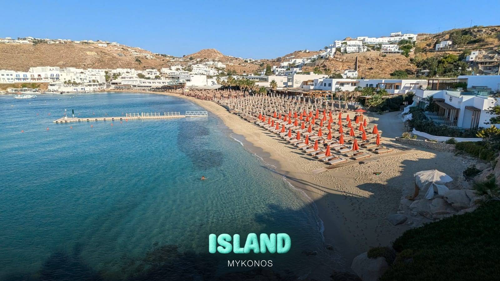 Platys Gialos beach on the south coast of Mykonos, with turquoise water, golden sand and rows of sunbeds, with the Party Island Mykonos logo